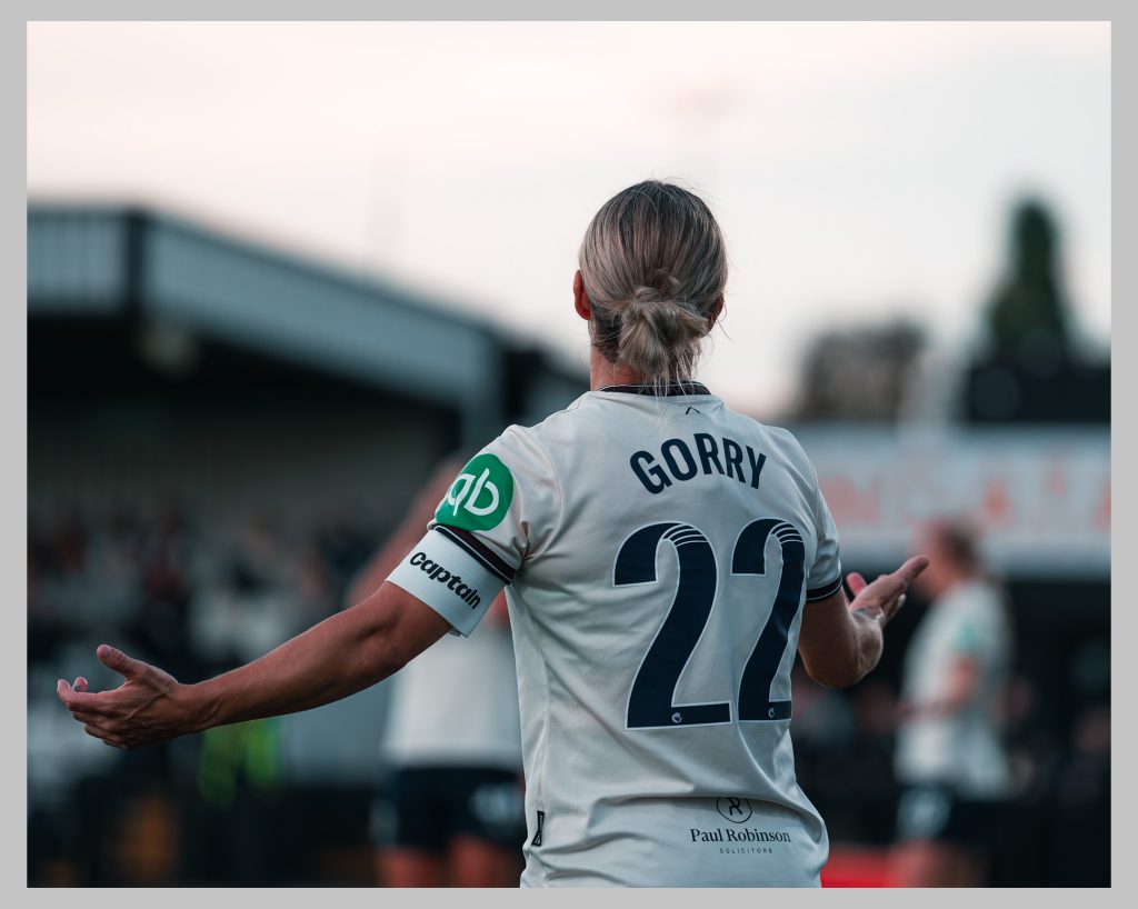 West Ham United captain Katrina Gorry pictured from behind in the club’s away kit, wearing number 22 and holding her arms out during a match. Text over the top reads: Gorry was a central figure during Australia’s run to the semi-finals of the 2023 Women’s World Cup, starting every match of the tournament. She later returned to the Olympic stage in Paris in 2024, having missed Tokyo while pregnant with Harper.
