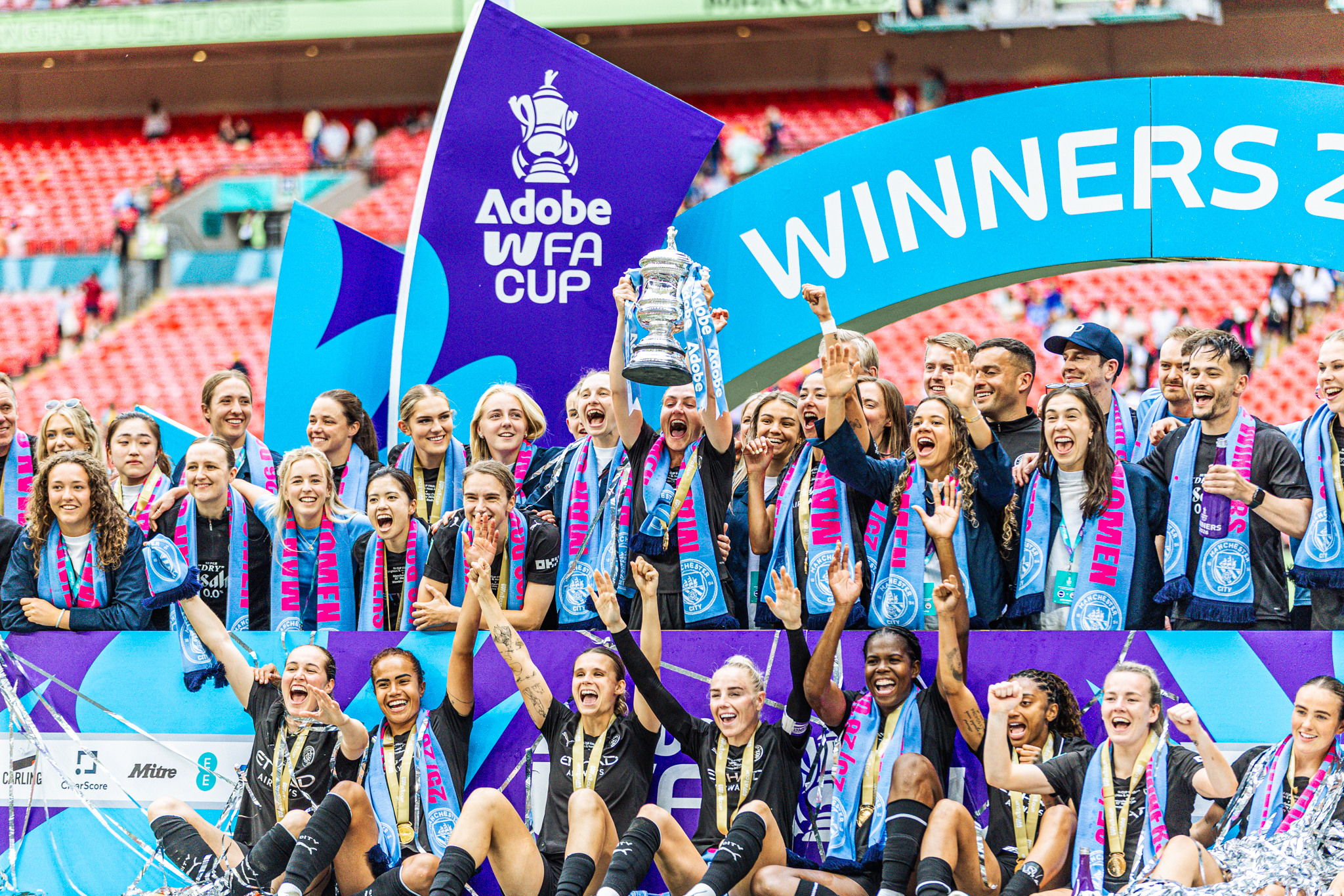 Manchester City Women celebrate at Wembley, lifting the Adobe Women’s FA Cup trophy beneath the winners’ arch as players and staff pose with their medals.