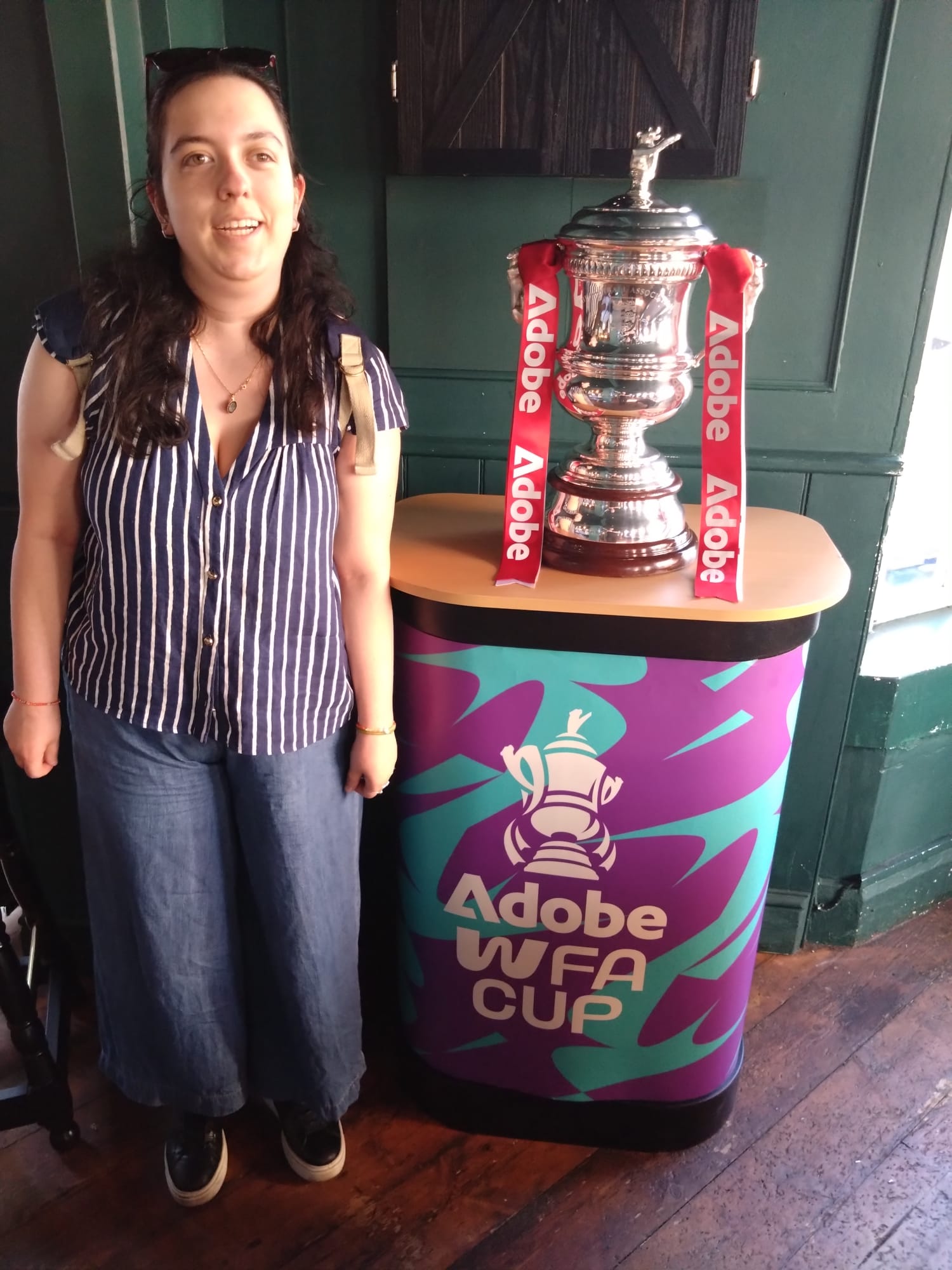 A person stands beside the Adobe Women's FA Cup trophy, displayed on a branded purple and turquoise pedestal with red Adobe ribbons. They are wearing a blue-and-white striped short-sleeved shirt, wide-leg blue jeans, black trainers and a backpack, smiling towards the camera in an indoor venue with dark green walls and wooden flooring.