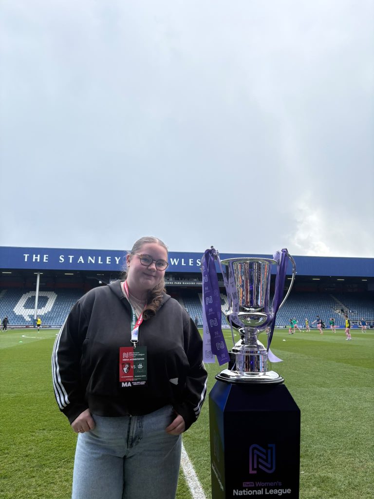 A person wearing a media accreditation pass stands beside the FA Women's National League trophy on the pitch at Loftus Road, home of Queens Park Rangers, before the National League Cup Final. Players warm up in the background beneath an overcast sky with a faint rainbow visible above the stadium.