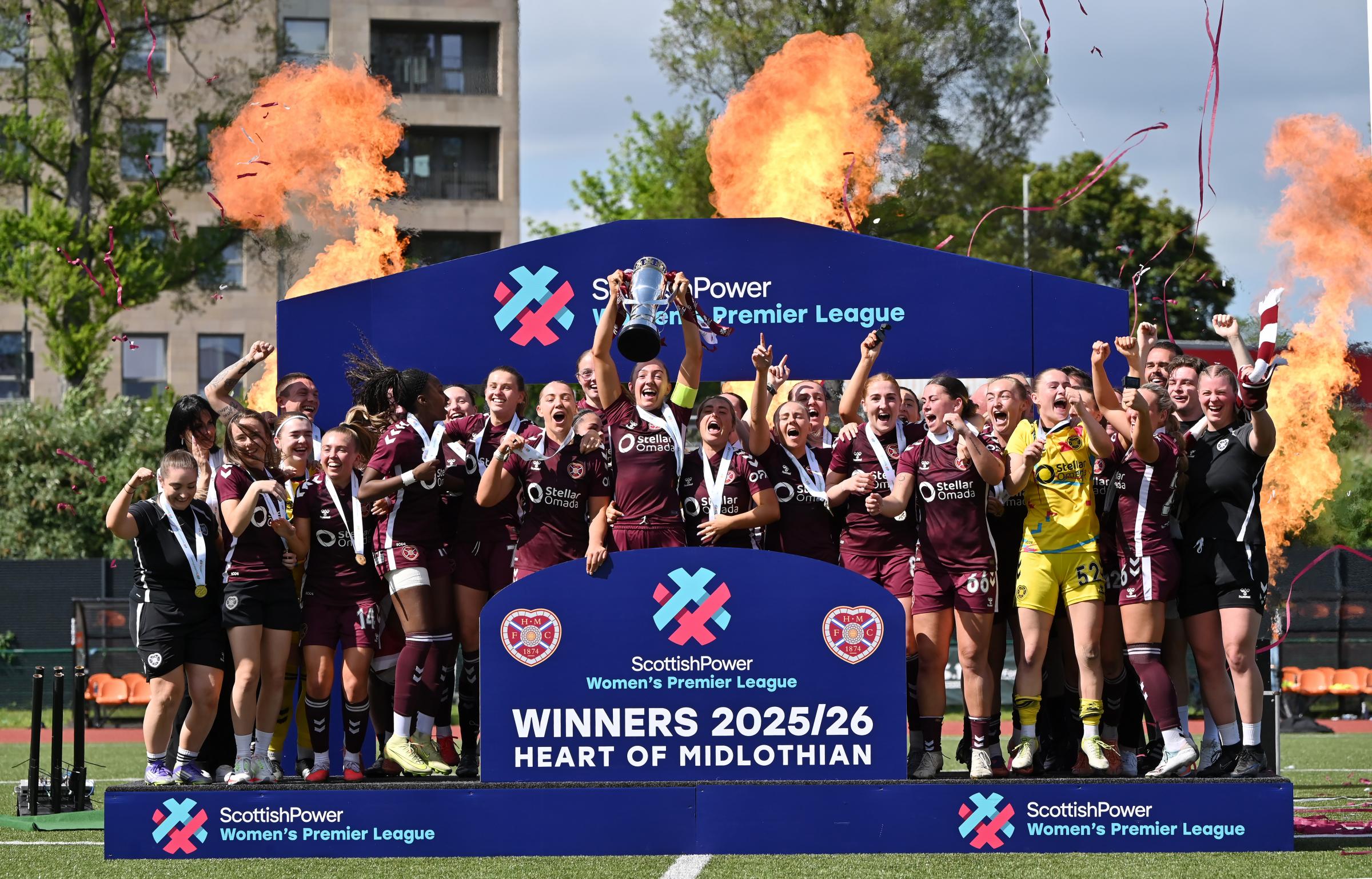 Heart of Midlothian Women celebrate winning the 2025/26 Scottish Power Women's Premier League title, with the captain lifting the trophy above her head on a winners' podium. Surrounded by jubilant teammates wearing medals, the squad cheers as confetti falls and pyrotechnic flames erupt behind the stage, with a sign reading "Winners 2025/26 – Heart of Midlothian.