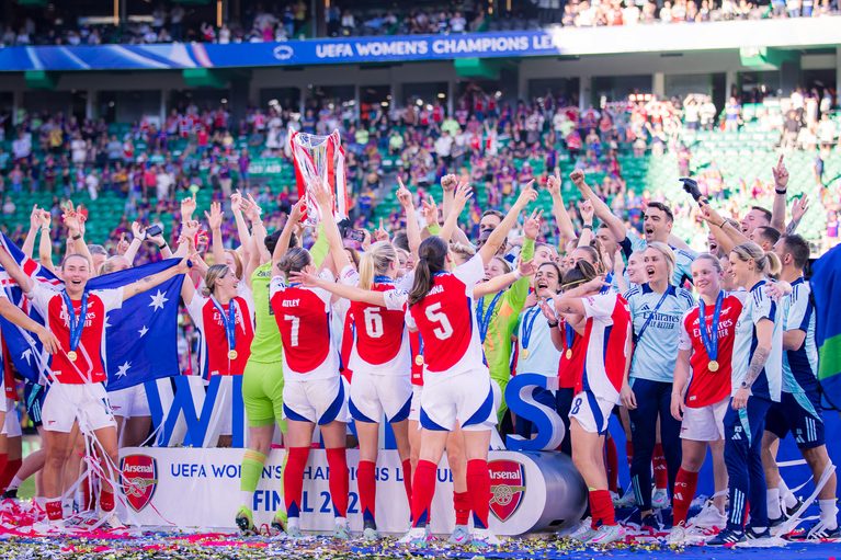 Arsenal Women players and staff celebrate together after winning the UEFA Women’s Champions League, gathered around the trophy with medals around their necks as confetti covers the pitch. An Australian flag is held aloft on the left, with supporters filling the stands behind them.