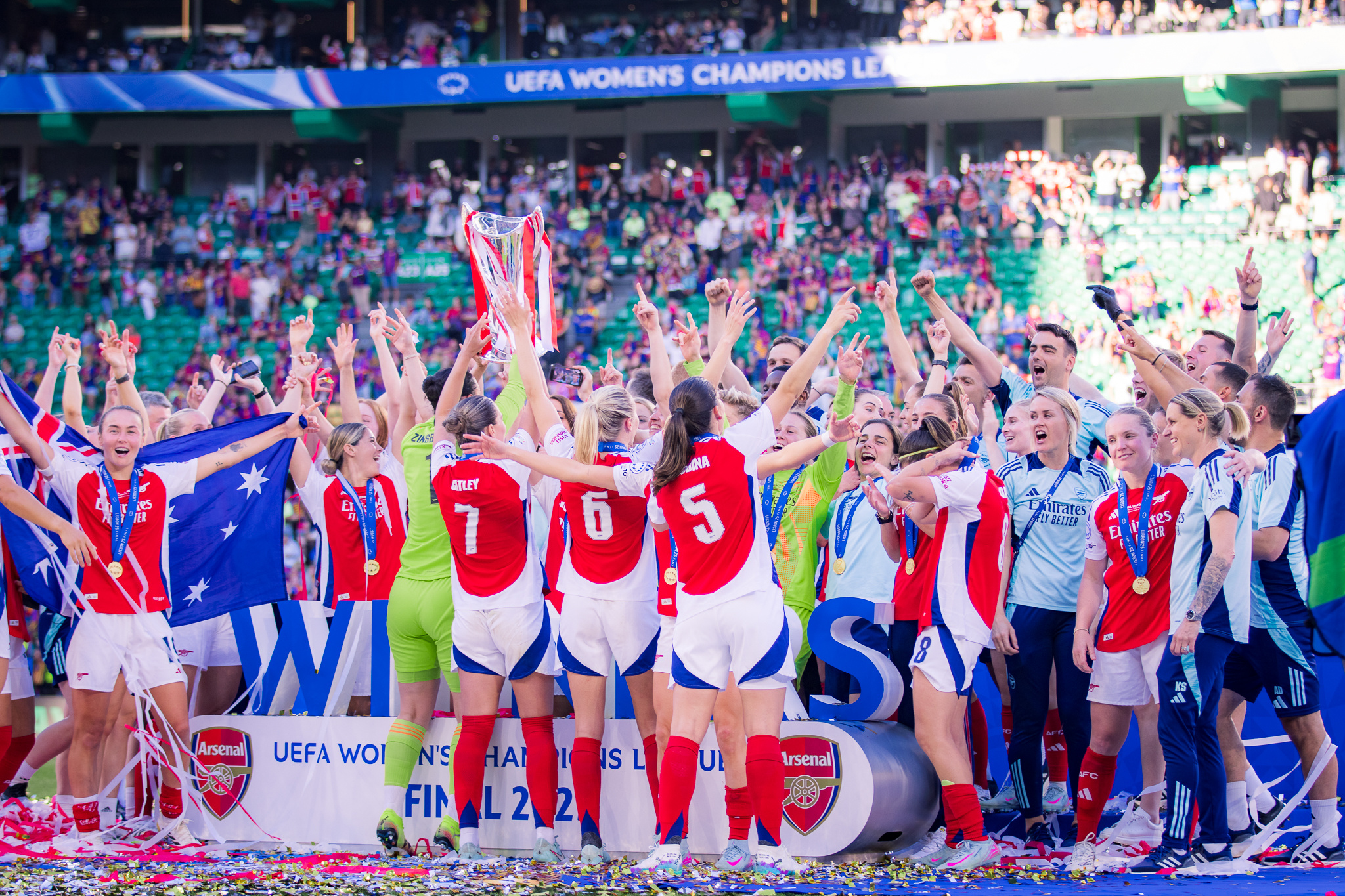 Arsenal Women players and staff celebrate together after winning the UEFA Women’s Champions League, gathered around the trophy with medals around their necks as confetti covers the pitch. An Australian flag is held aloft on the left, with supporters filling the stands behind them.