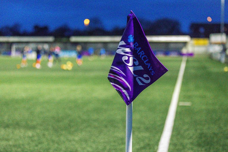 A Barclays WSL 2 corner flag beside the pitch, with players warming up in the blurred background under an evening sky.