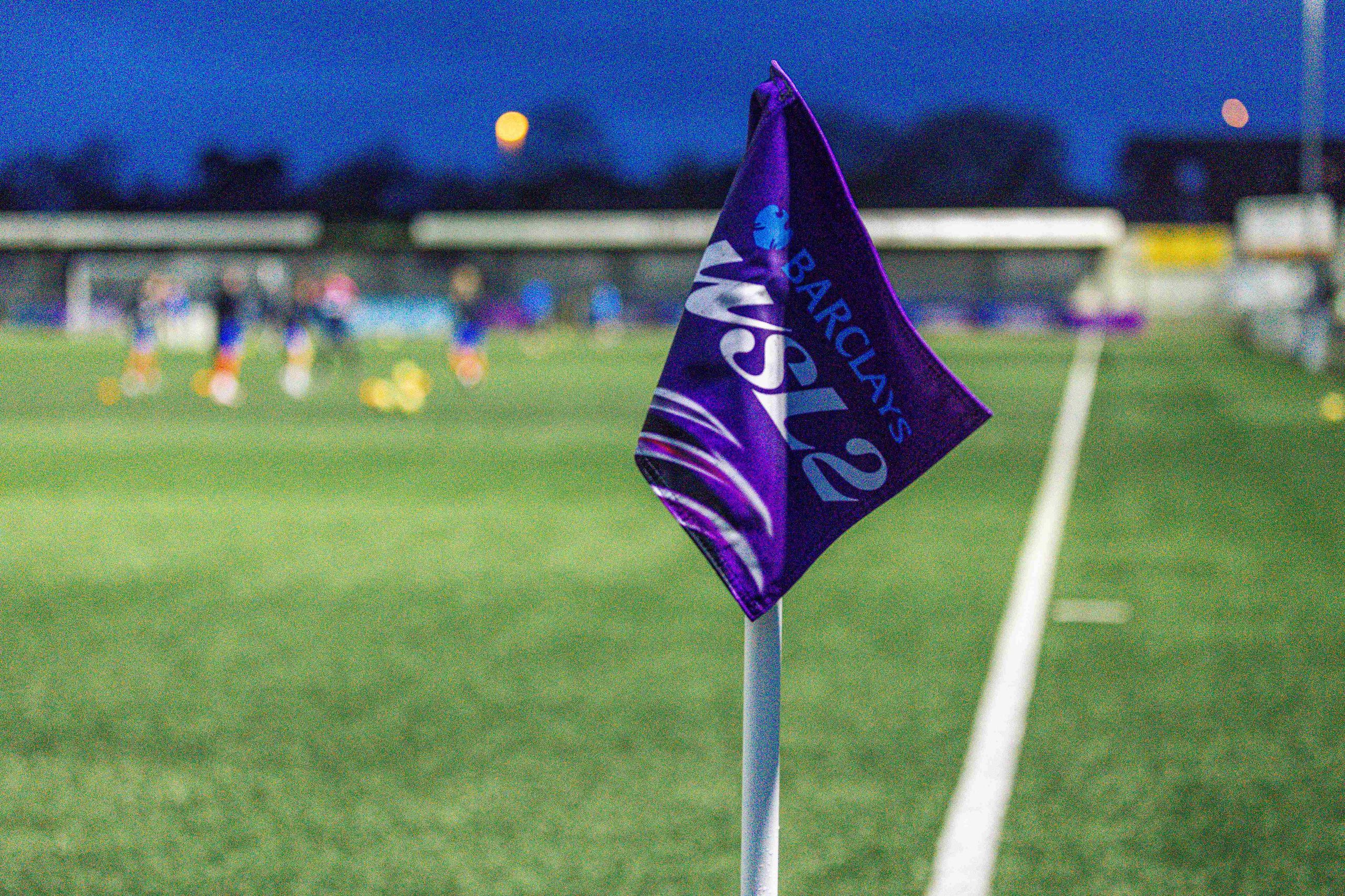 A Barclays WSL 2 corner flag beside the pitch, with players warming up in the blurred background under an evening sky.