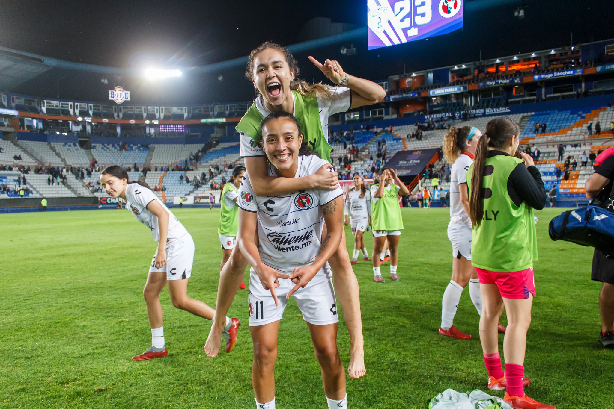 Two Club Tijuana Femenil players celebrate on the pitch after a match, with one jumping onto her teammate’s back as they smile towards the camera.