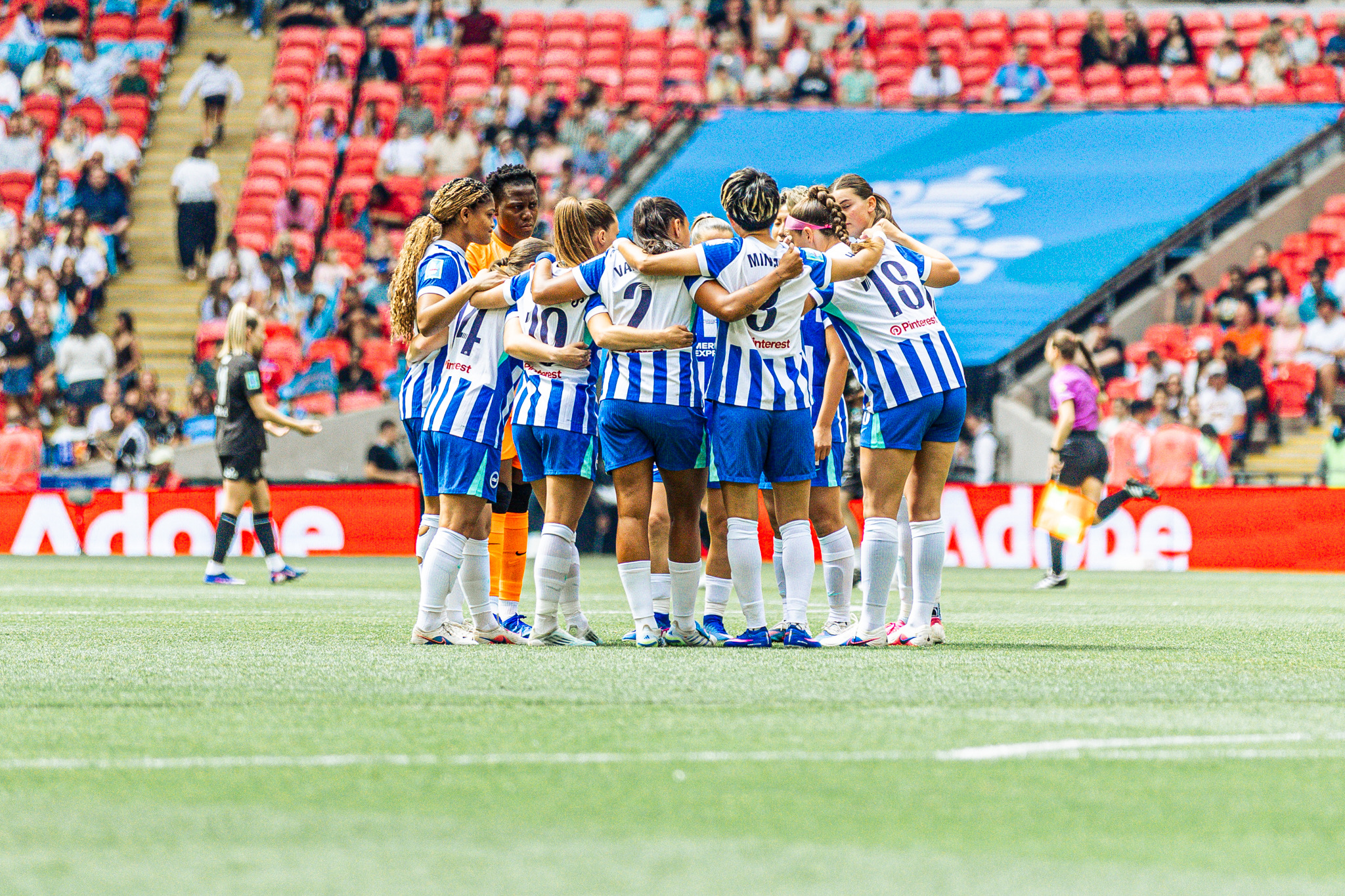 Brighton & Hove Albion Women players huddle together on the pitch before a match, wearing blue-and-white striped shirts, with red stadium seats and spectators in the background.