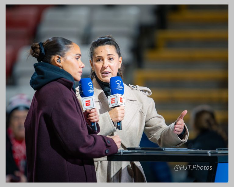 Fara Williams and Alex Scott as football pundits stand pitchside holding ESPN-branded UEFA Women’s Champions League microphones. One wears a dark burgundy coat, while the other wears a light beige coat and gestures with her hand as she speaks. Stadium seating is blurred in the background.