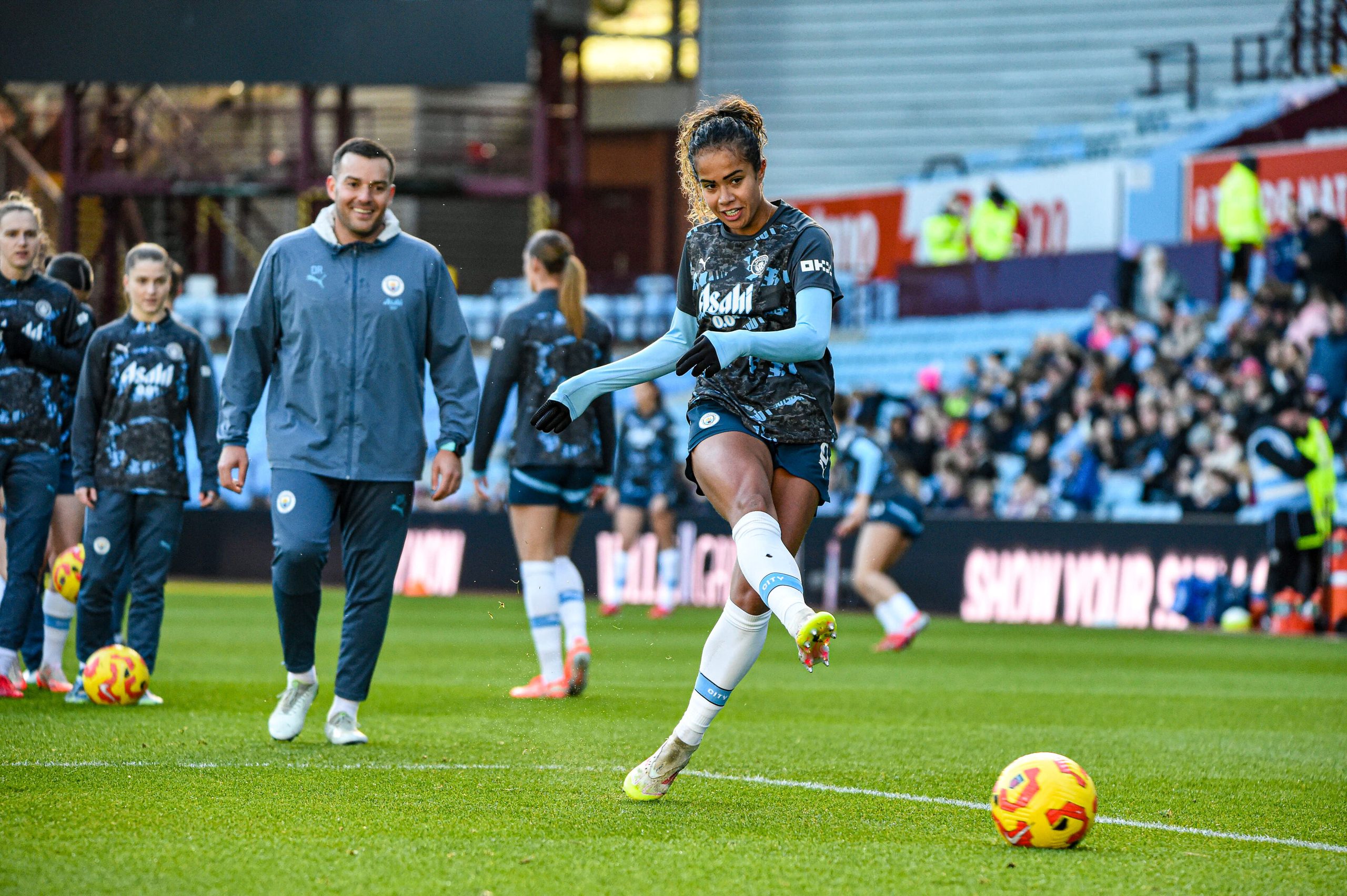 Mary Fowler strikes a ball during the warm-up before a match, wearing the club’s dark patterned training shirt and light blue undershirt. Teammates and coaching staff are visible behind her on the pitch, with supporters in the stands in the background.