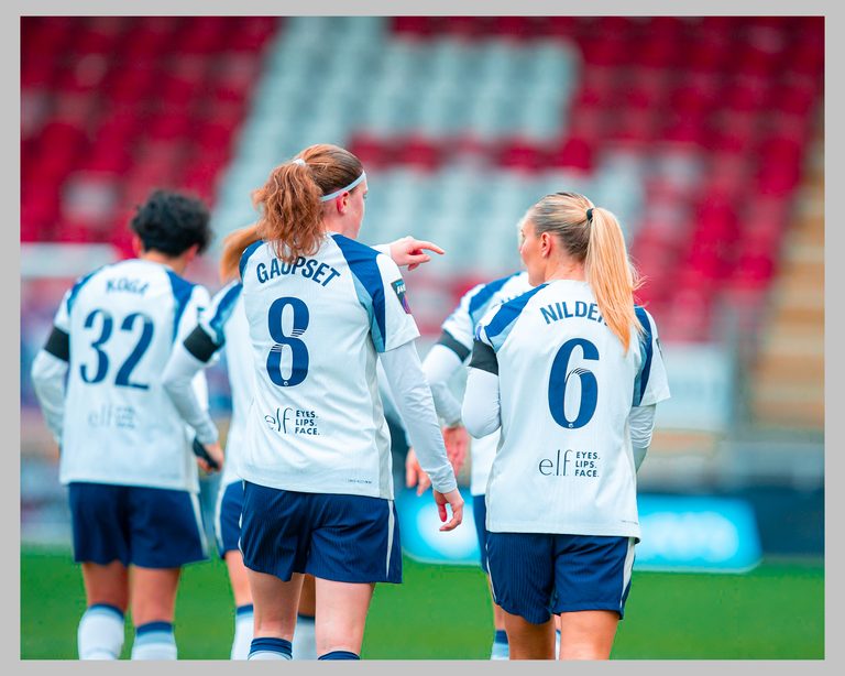 Spurs players Gaupset and Nilden walk together on the pitch, seen from behind in their white and blue kits, with teammates and red stadium seats in the background.