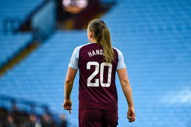 Kirsty Hanson wearing a claret and blue kit with “Hanson” and the number 20 on the back, walking across the pitch with rows of blue stadium seats in the background.