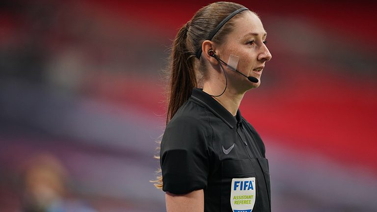 Sian Massey-Ellis wearing a black FIFA uniform and headset, pictured pitchside at a football stadium.