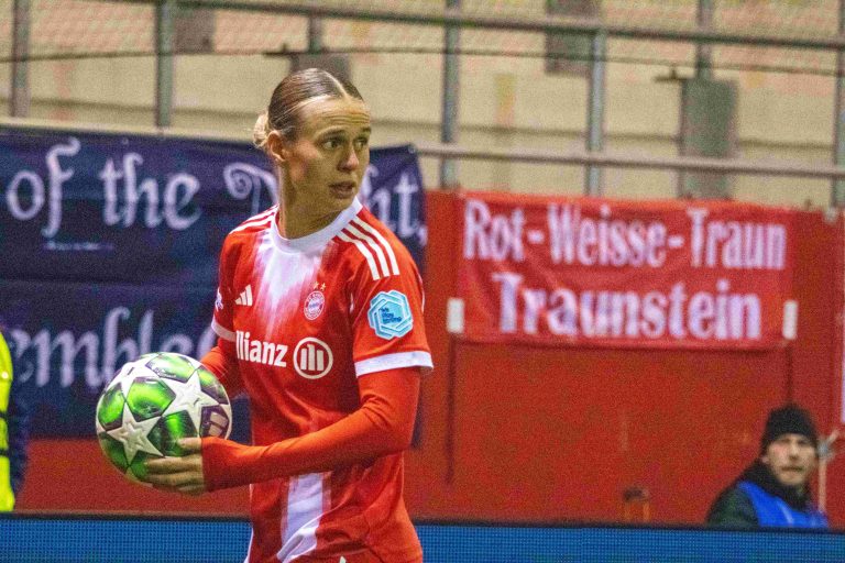 Klara Bühl holds the ball on the touchline while wearing Bayern Munich’s red home kit during a match. A red Bayern supporters’ banner is visible in the stands behind her.