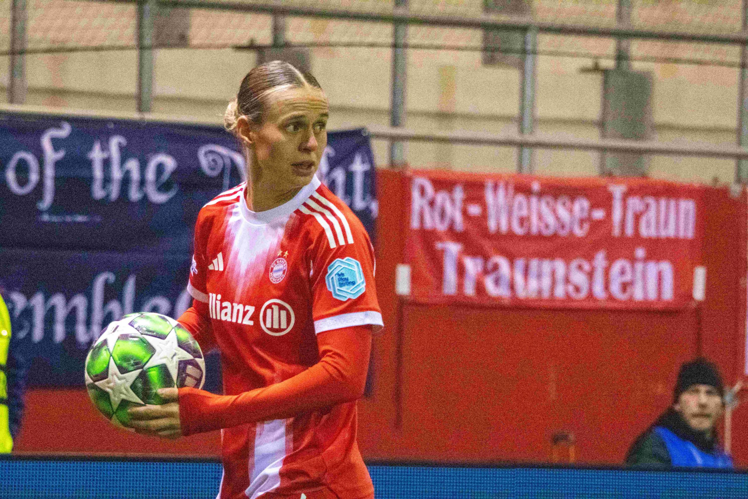 Klara Bühl holds the ball on the touchline while wearing Bayern Munich’s red home kit during a match. A red Bayern supporters’ banner is visible in the stands behind her.