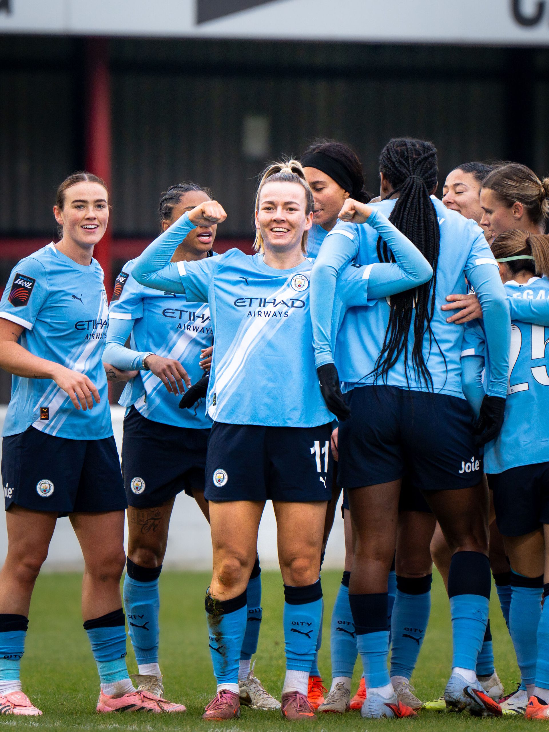 Lauren Hemp celebrates for Manchester City with both arms raised and fists clenched, smiling as her teammates gather around her on the pitch.