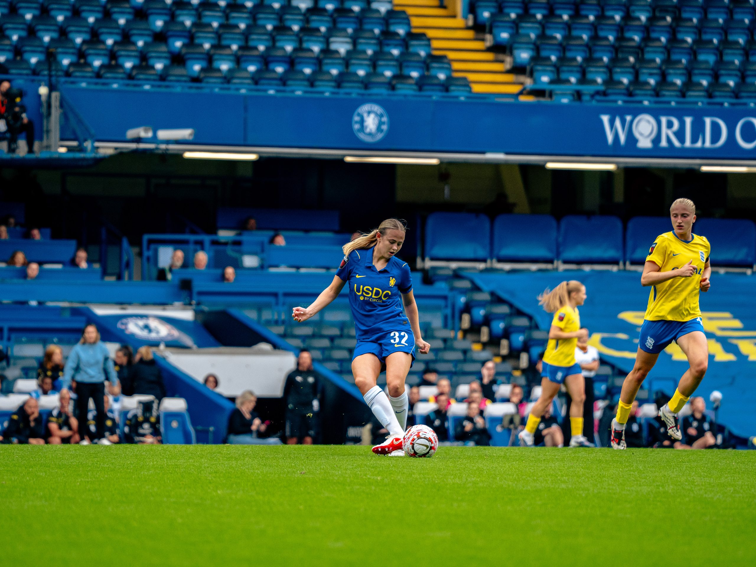 Lexi Potter controls the ball on the pitch at Stamford Bridge during a match, with two opposition players in yellow and blue kits nearby. Blue stadium seating and Chelsea branding are visible in the background.