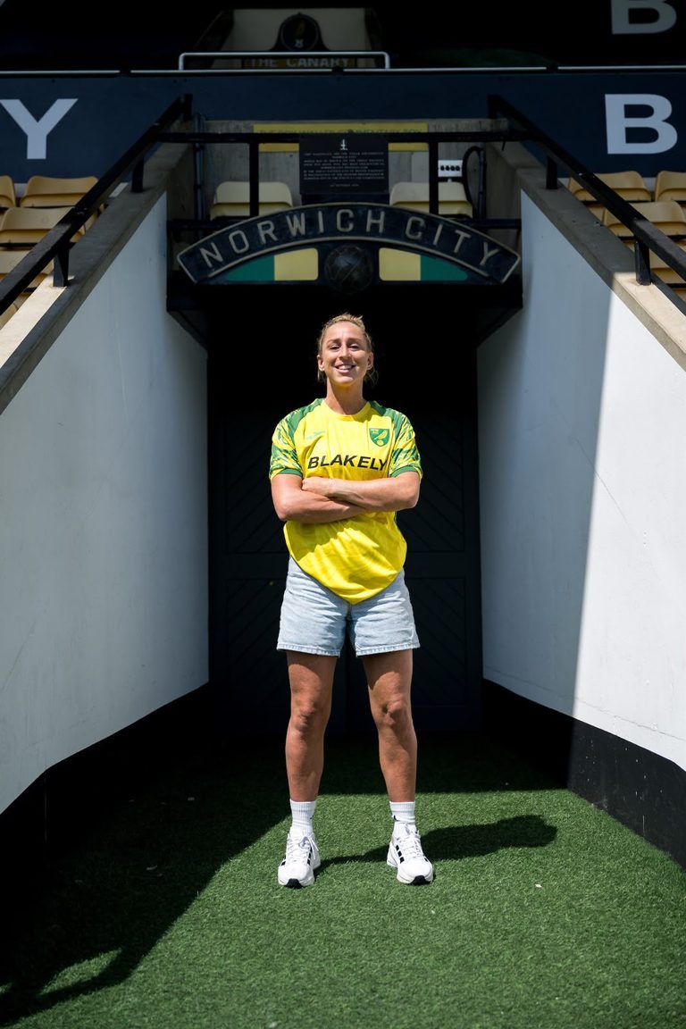 Natalie Johnson wearing a yellow and green Norwich City shirt stands with her arms crossed in the players’ tunnel at Carrow Road, beneath a Norwich City sign.