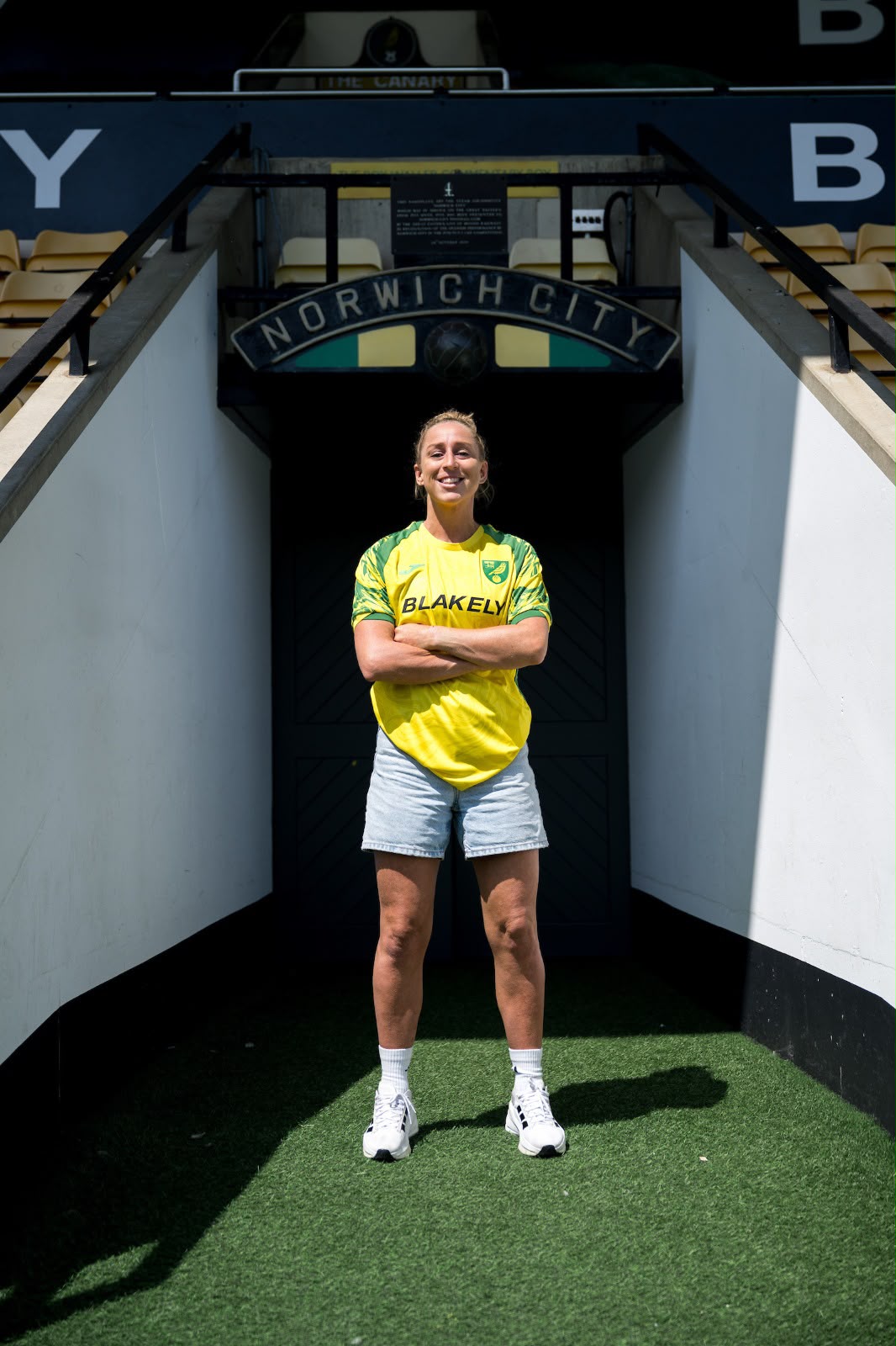 Natalie Johnson wearing a yellow and green Norwich City shirt stands with her arms crossed in the players’ tunnel at Carrow Road, beneath a Norwich City sign.