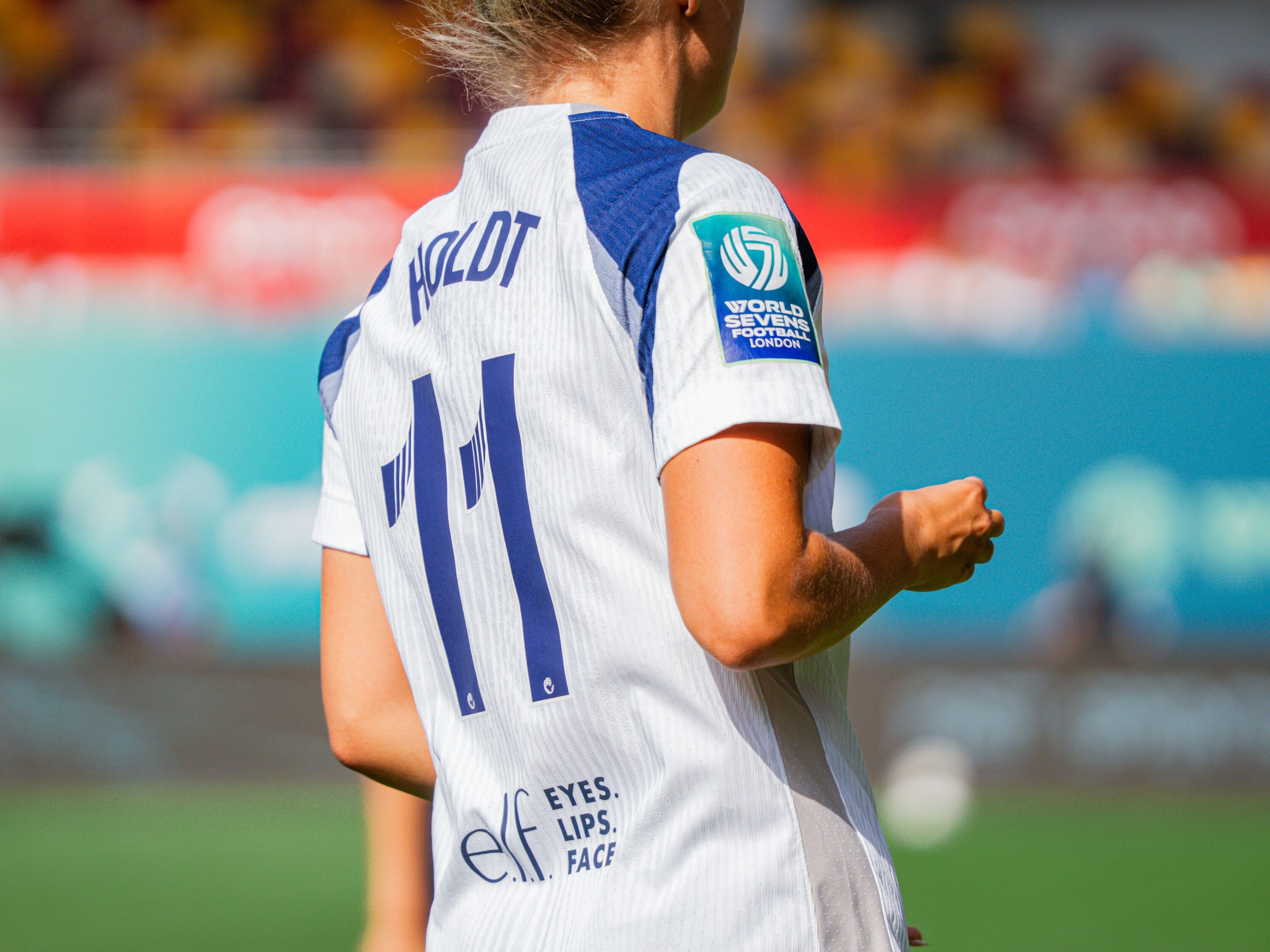 Close-up rear view of a footballer wearing a white shirt with “Holdt” and the number 11 on the back, featuring a World Sevens Football London sleeve patch.