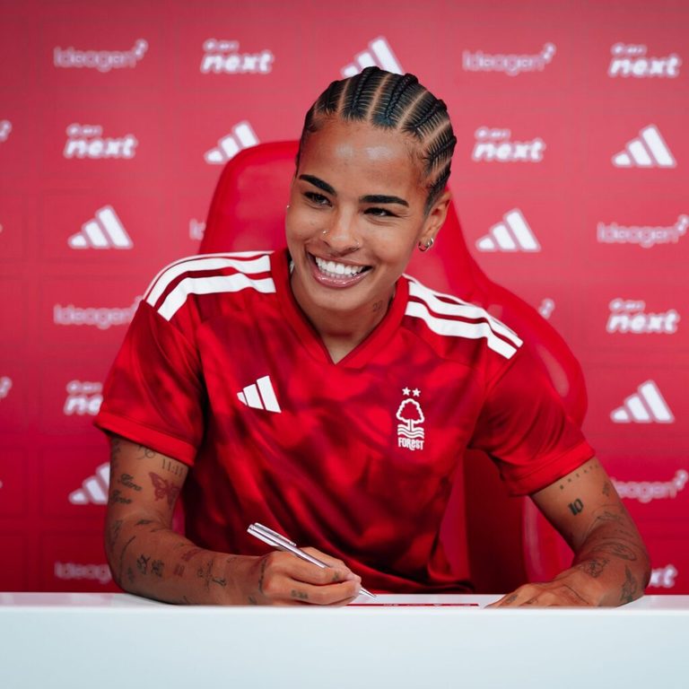 Shania Hayles sits at a desk signing a document while wearing the club’s red Adidas shirt, with a red sponsor backdrop behind her.