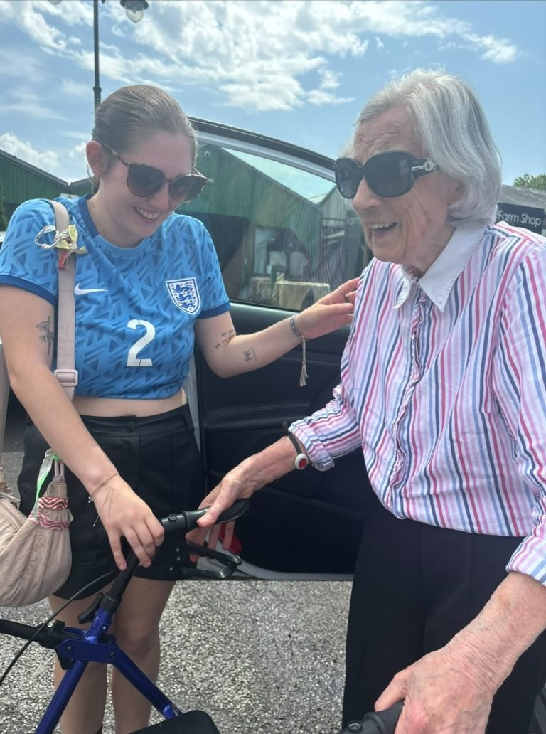 Two women smile together beside an open car door on a sunny day. A younger woman wearing a blue England football shirt stands alongside an older woman wearing a striped shirt and sunglasses, who is holding onto a walking frame.