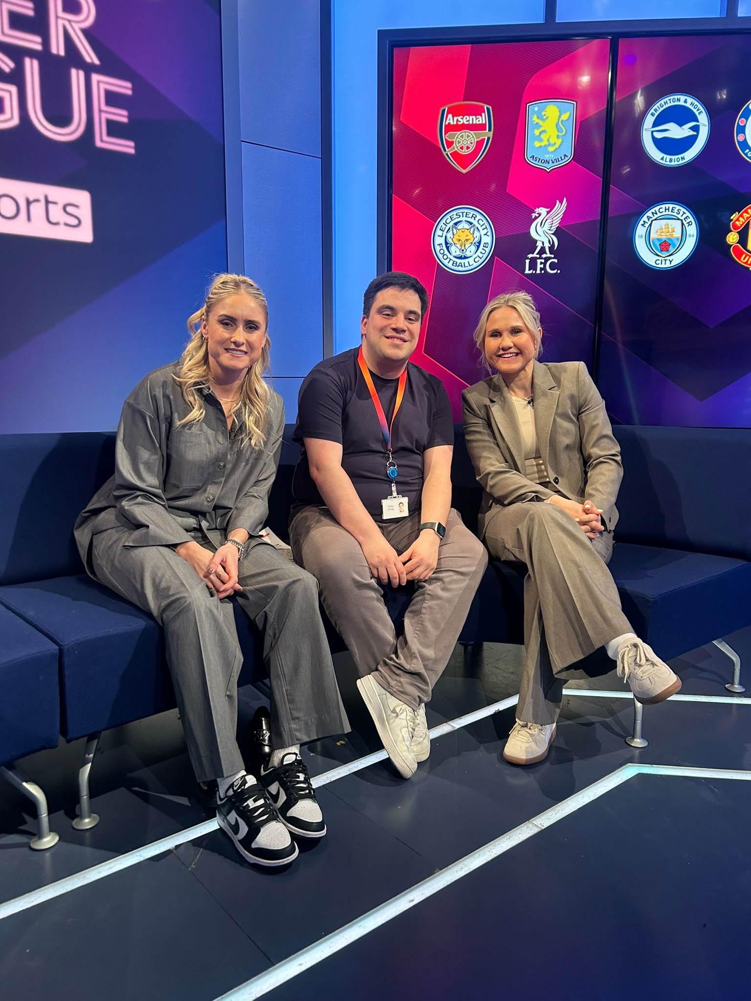 Three people smiling while seated together on a sofa in a TV sports studio, with a screen displaying Women’s Super League club badges behind them.