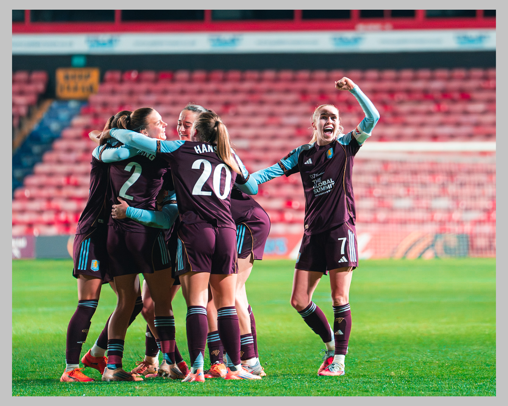 Aston Villa players celebrate together on the pitch, with several players embracing in a group while a teammate raises her fist in celebration beside them.