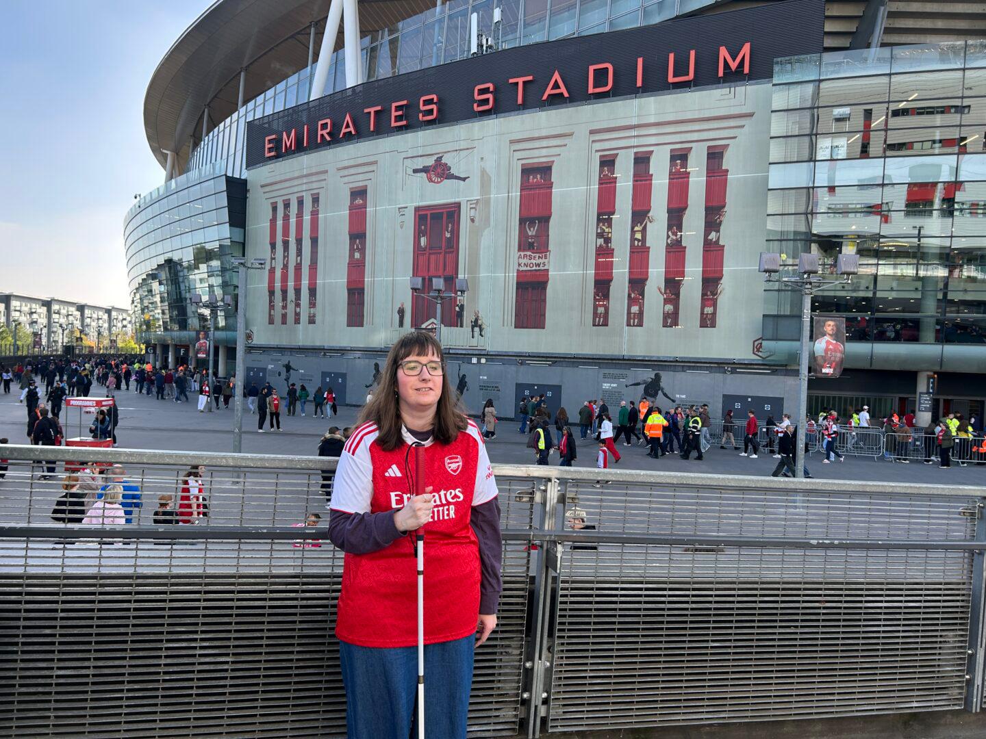 A woman wearing an Arsenal home shirt stands outside Emirates Stadium, holding a white cane. The stadium façade and large “EMIRATES STADIUM” sign are visible behind her, with supporters gathering around the entrances before a match.