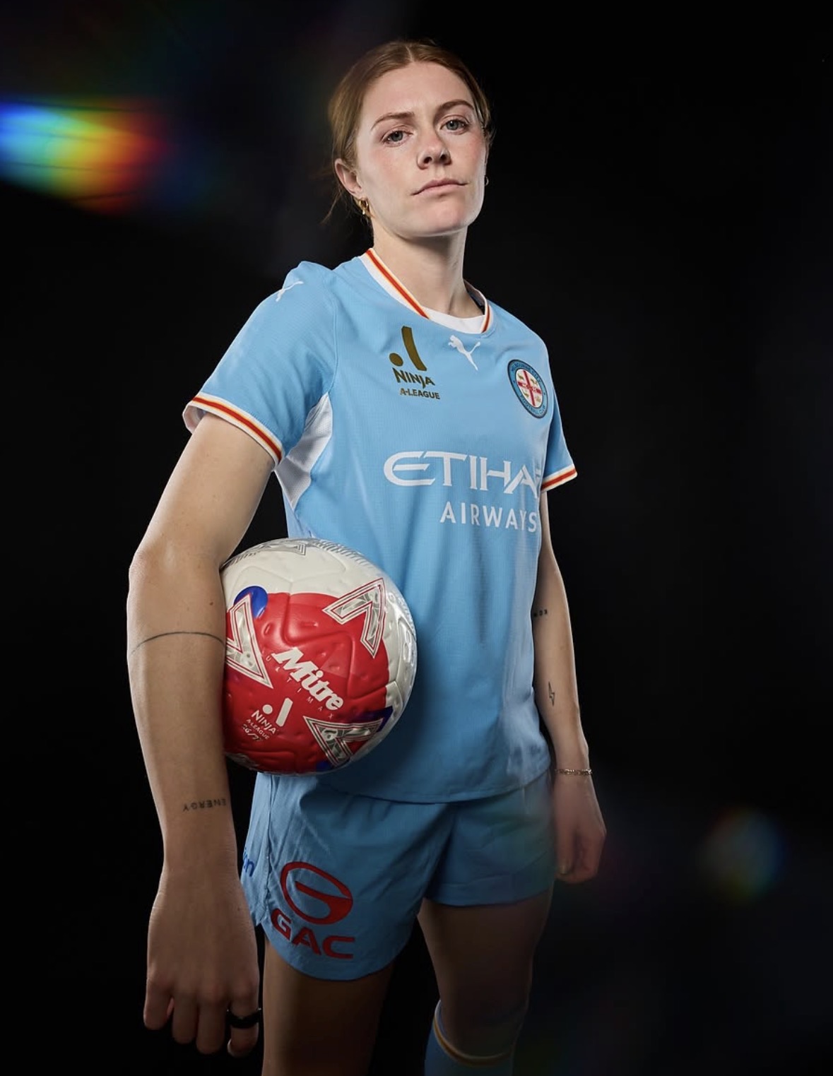 Cortnee Vine poses against a black background in Melbourne City’s sky-blue kit, holding a red and white Ninja A-League football under one arm. A rainbow light effect appears to the left of the image.