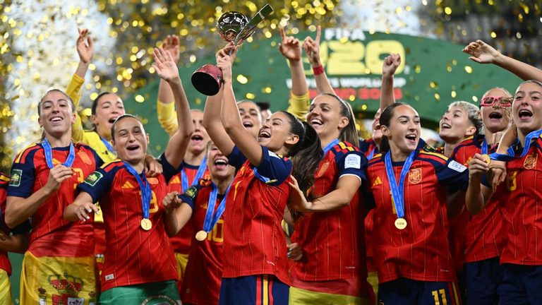 Spain players celebrate with gold medals as a teammate lifts the FIFA U-20 Women’s World Cup trophy above her head, surrounded by falling gold confetti.