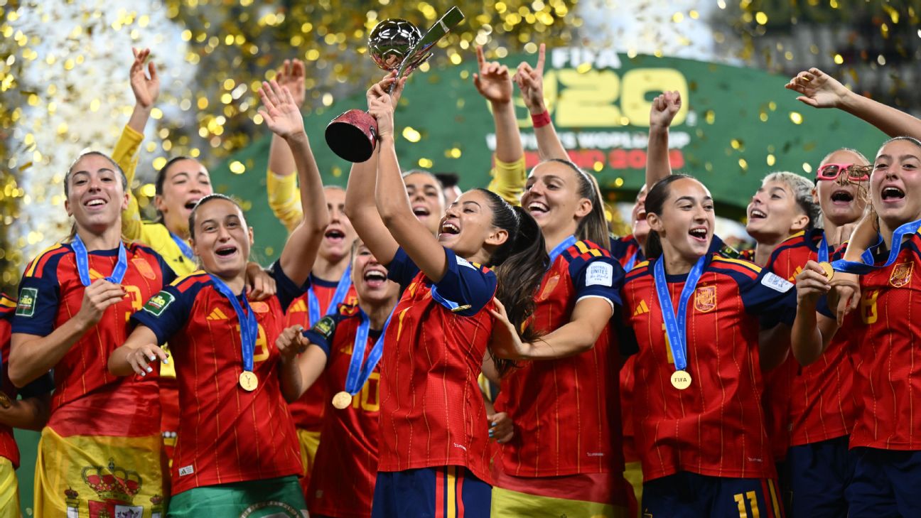 Spain players celebrate with gold medals as a teammate lifts the FIFA U-20 Women’s World Cup trophy above her head, surrounded by falling gold confetti.